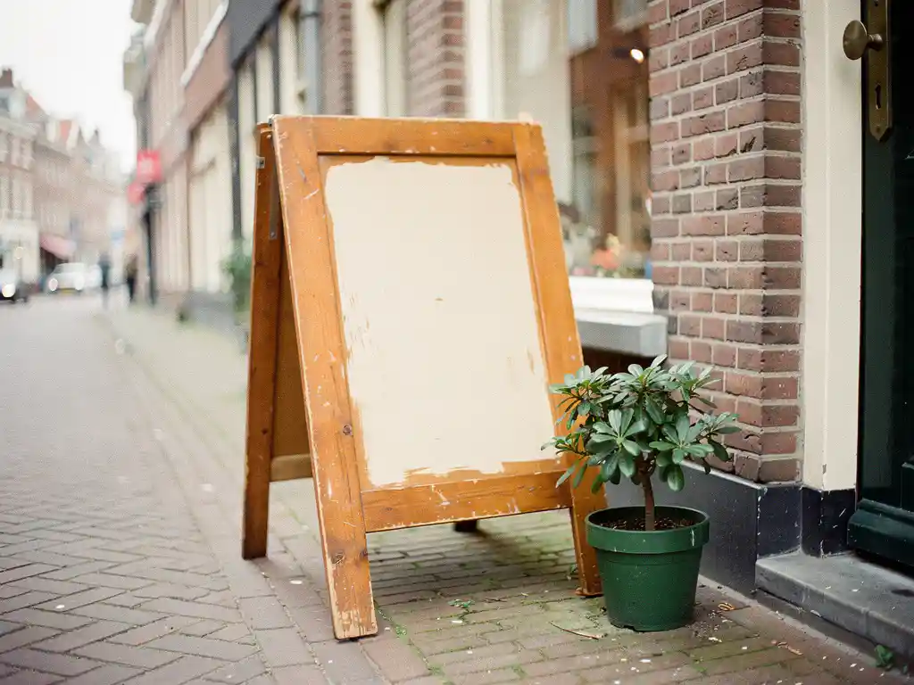 Handgeschilderd houten sandwichbord tegen bakstenen gevel op Nederlandse straat, met klein potplantje ernaast in zacht daglicht.