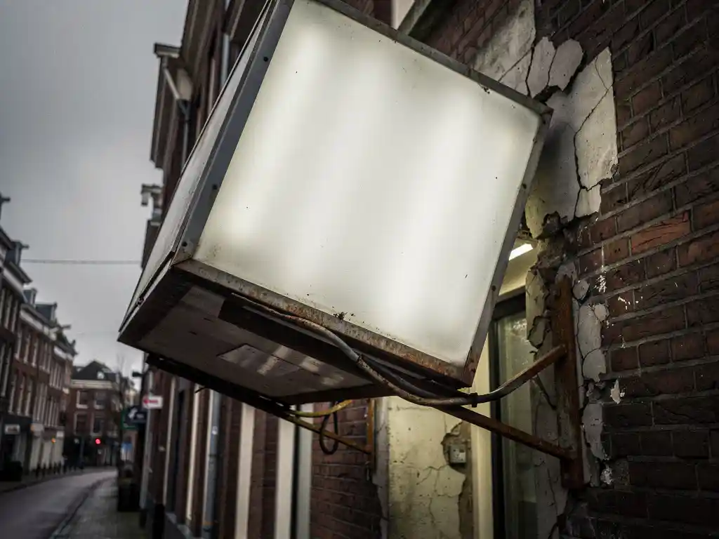 Loszittend verlicht reclamebord dat van een bakstenen gevel aftrekt, met verbogen beugels en afbrokkelend voegwerk op een bewolkte Nederlandse straat.