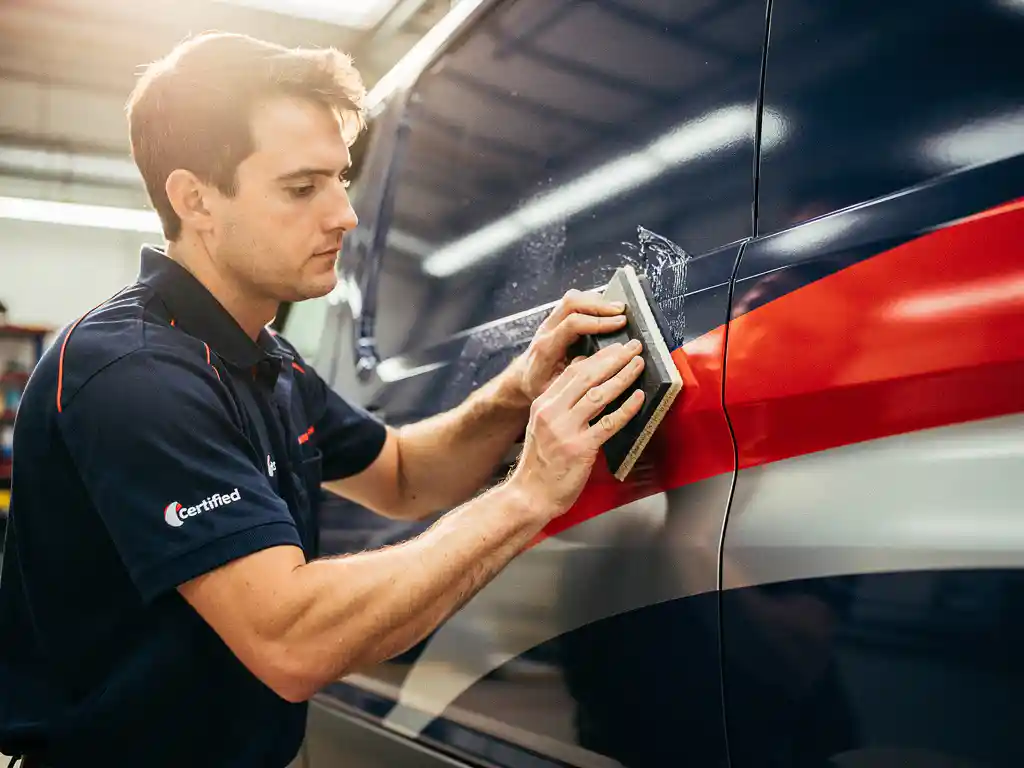Gecertificeerde technicus brengt vinyl voertuigwrap aan op bestelwagen met squeegee, professionele werkplaats met felle verlichting.